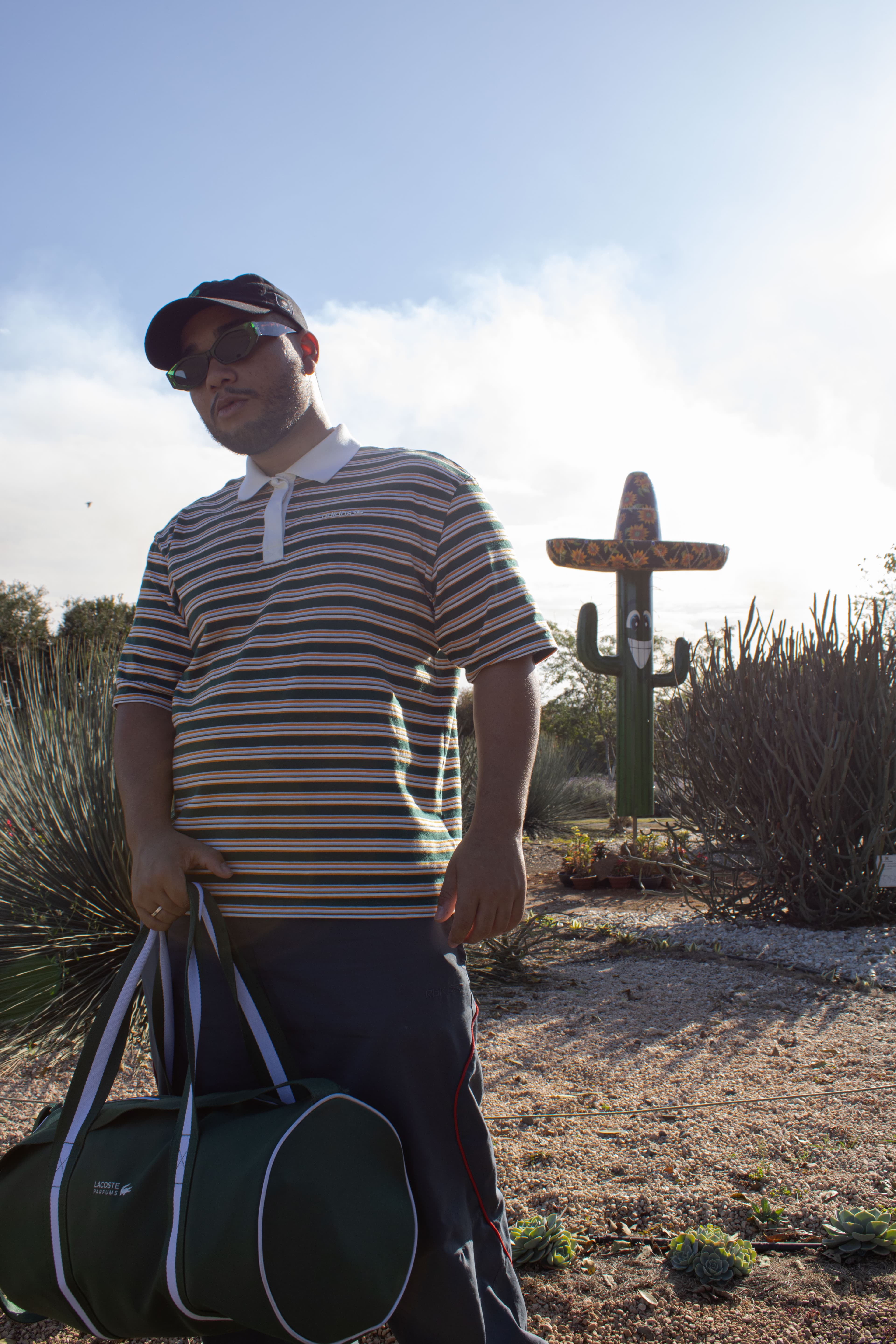 Retrato com a proposta do Antigo México, o modelo com expressão séria, segura uma maleta no deserto com um cacto com chapéu mexicano e com um sorriso.